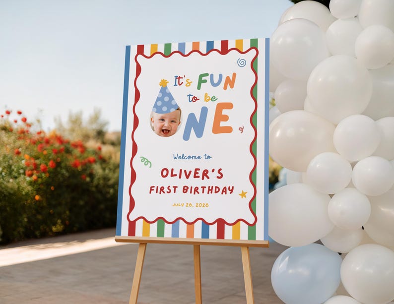 May include: A rectangular birthday sign on a wooden easel featuring a colorful striped border, a photo of a child in a blue party hat, and the text: It's FUN to be ONE, Welcome to OLIVER'S FIRST BIRTHDAY, JULY 26, 2026, with small graphic accents.