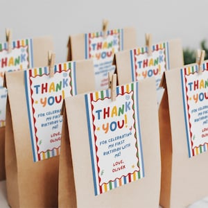 May include: A group of brown paper bags, each featuring a rectangular tag attached with a wooden clip. The tags have a colorful striped border and the text: THANK YOU! FOR CELEBRATING MY FIRST BIRTHDAY WITH ME! LOVE, OLIVER, in multi-colored letters.