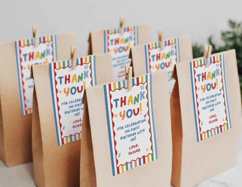 May include: A group of brown paper bags, each featuring a rectangular tag attached with a wooden clip. The tags have a colorful striped border and the text: THANK YOU! FOR CELEBRATING MY FIRST BIRTHDAY WITH ME! LOVE, OLIVER, in multi-colored letters.