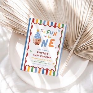 May include: A colorful first birthday invitation with the text "It's FUN to be ONE." The invitation features a photo of a baby wearing a party hat and includes details for Oliver's first birthday celebration. The design has a striped border with red, orange, yellow, green, and blue.