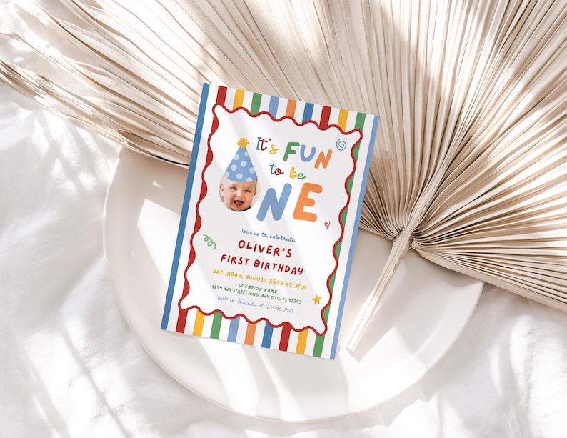 May include: A colorful first birthday invitation with the text "It's FUN to be ONE." The invitation features a photo of a baby wearing a party hat and includes details for Oliver's first birthday celebration. The design has a striped border with red, orange, yellow, green, and blue.