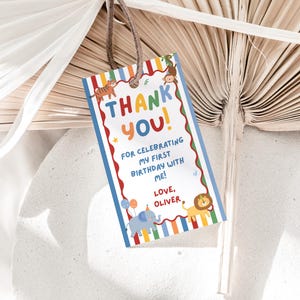 Puede incluir: Una etiqueta de regalo rectangular con un centro blanco y un borde de rayas coloridas. Presenta animales de dibujos animados que incluyen un tigre, un mono, un elefante y un león. El texto dice: THANK YOU! FOR CELEBRATING MY FIRST BIRTHDAY WITH ME! LOVE, OLIVER.