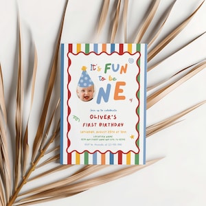 May include: A colorful birthday invitation with the text "It's FUN to be ONE." The invitation features a photo of a baby wearing a party hat and is decorated with a striped border in various colors. The invitation is for Oliver's first birthday.