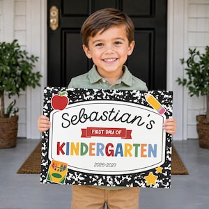 May include: A rectangular sign with a black and white composition notebook pattern border, featuring the text "Sebastian's FIRST DAY OF KINDERGARTEN 2026-2027" with illustrations of a red apple, a yellow pencil, a box of crayons, and a yellow star.