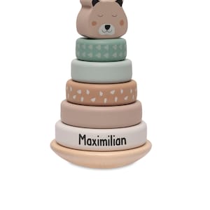Könnte beinhalten: Ein Holz-Steckspielzeug mit einem Bärenkopf und sechs farbigen Ringen. Die Ringe sind in Rosa, Grün und Beige gehalten, mit dem Namen "Maximilian" auf der Basis. Der Bär hat ein süßes Gesicht mit geschlossenen Augen.