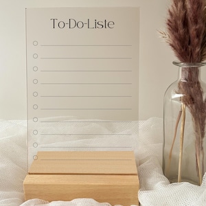Może przedstawiać: Przezroczysty akrylowy notatnik To-Do-Liste z drewnianą podstawą. Notatnik ma listę linii z kółkami do zaznaczania. Tekst "To-Do-Liste" jest wydrukowany na czarno. Po prawej stronie znajduje się przezroczysty szklany wazon z suszonymi kwiatami.