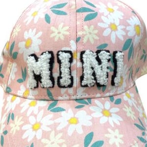 May include: Pink baseball cap with a daisy floral pattern and the word "MINI" in white, textured letters. The cap has a curved brim and a rounded crown. The hat is designed for children.