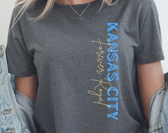 Może przedstawiać: Grafitowy t-shirt z pionowym nadrukiem "KANSAS CITY forever Royal" w kolorze niebieskim i złotym. Koszulka ma krótkie rękawy i jest noszona z dżinsową kurtką.