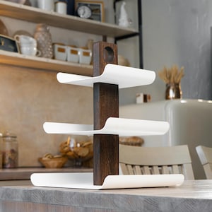 Könnte beinhalten: Ein weißer, mehrstufiger Servierständer mit einem dunkelbraunen Holzpfosten in der Mitte. Der Ständer hat drei Ebenen und ist aus Metall. Er steht auf einer hellbraunen Arbeitsplatte in einer Küche.