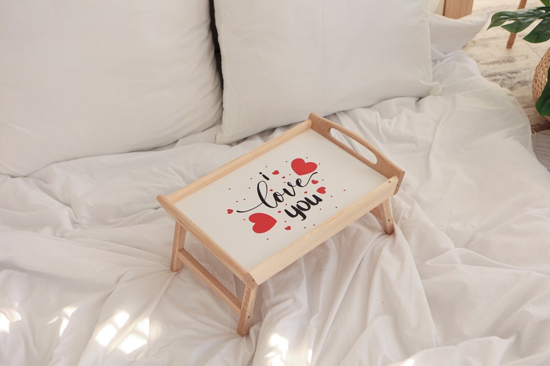 Klappbarer Betttisch aus Holz mit romantischem Herzmotiv auf einem weißen Sofa, mit ausgeklappten Holzbeinen.