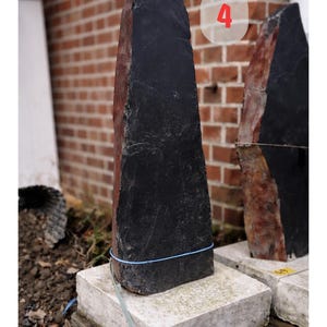 May include: A tall, dark gray stone sculpture with a rough texture and a reddish-brown side. The stone is approximately 31.5 x 12.6 x 3.1 inches and rests on a concrete base. A blue string wraps around the stone.