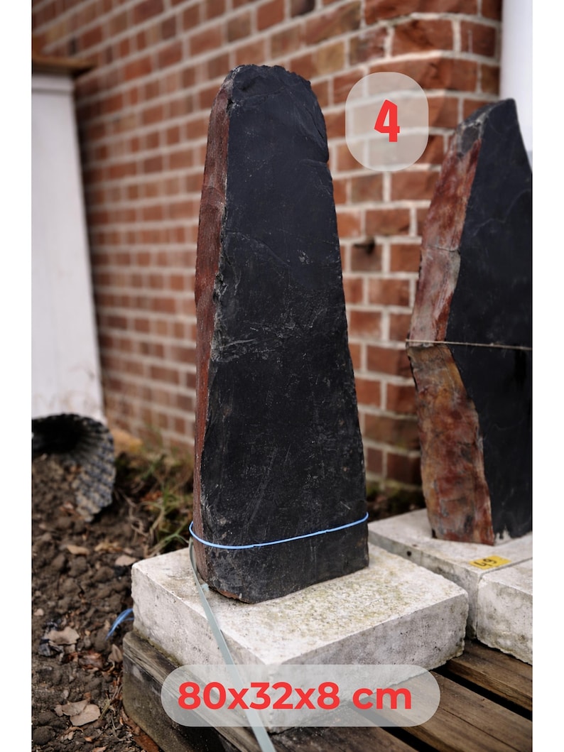 May include: A tall, dark gray stone sculpture with a rough texture and a reddish-brown side. The stone is approximately 31.5 x 12.6 x 3.1 inches and rests on a concrete base. A blue string wraps around the stone.