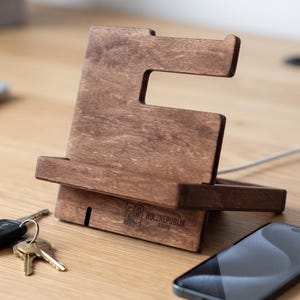 Könnte beinhalten: Ein dunkelbrauner Handyhalter aus Holz mit einer Aussparung und einer horizontalen Ablage für das Gerät. Auf der Basis befindet sich die Gravur HOLZREPUBLIK STADE mit einem kleinen Haussymbol. Ein Ladekabel ist an der Rückseite sichtbar.