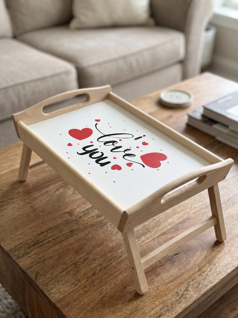 Klappbares Frühstückstablett aus Holz mit weißer Platte, roten Herzen und Schriftzug auf einem Holztisch.