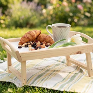 Frühstückstablett aus hellem Holz mit klappbaren Beinen auf einer Picknickdecke im Garten, dekoriert mit Tasse und Gebäck.