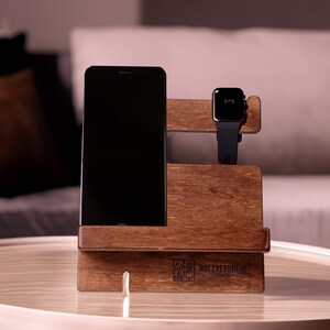Könnte beinhalten: Ein Holzständer für Telefon und Uhr mit dunkelbrauner Oberfläche. Der Ständer hat einen Schlitz für ein Telefon und einen Platz für eine Uhr. Der Ständer ist mit dem Text "HOLZREPUBLIK STADE" graviert.