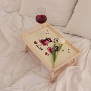 Frühstückstablett aus Holz mit rosa Blumenmotiv und klappbaren Beinen auf einem hellen Sofa.