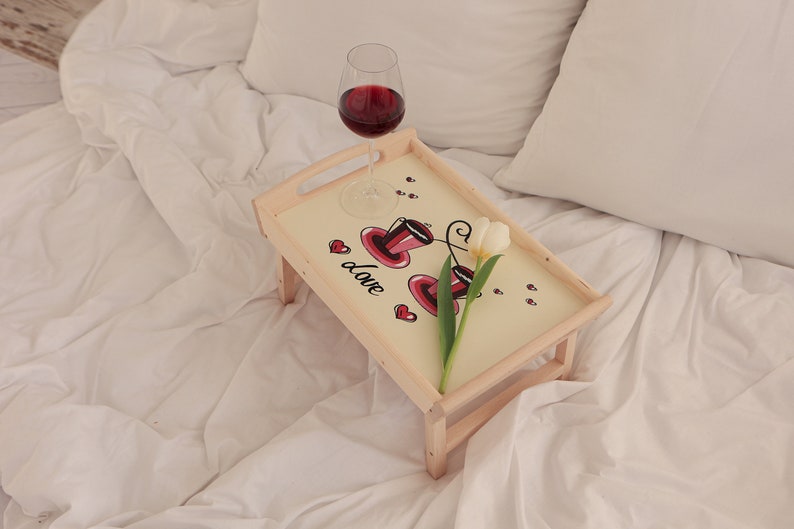 Frühstückstablett aus Holz mit rosa Blumenmotiv und klappbaren Beinen auf einem hellen Sofa.