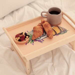 Serviertablett aus Holz mit Bienenmotiv, Kaffee, Croissant und Obst auf einer hellen Bettdecke.