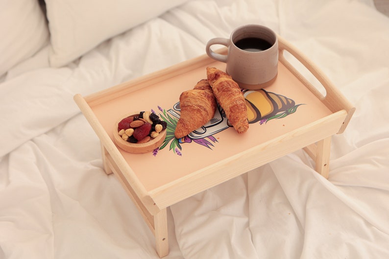 Serviertablett aus Holz mit Bienenmotiv, Kaffee, Croissant und Obst auf einer hellen Bettdecke.