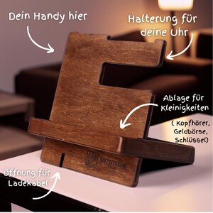 Könnte beinhalten: Ein Holz-Telefonständer mit einem Schlitz für ein Telefon, einem kleinen Regal für Ladekabel und einem separaten Regal für eine Uhr oder andere Kleinigkeiten. Der Ständer ist mit "HOLZREPUBLIK" beschriftet und trägt den Text "Dein Handy hier", "Halterung für deine Uhr", "Öffnung für Ladekabel", "Ablage für Kleinigkeiten (Kopfhörer, Geldbörse, Schlüssel)".