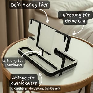 Könnte beinhalten: Ein weißer und schwarzer Holz-Telefonständer mit einem Schlitz für ein Telefon, einem kleinen Regal für Ladekabel und einem separaten Regal für Kleinigkeiten. Der Text "Dein Handy hier" ist auf dem Ständer geschrieben.