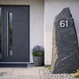 May include: A dark gray stone address marker with the number 61 in silver. The marker stands in front of a dark gray door and a white wall. Potted plants and a doormat are also visible.