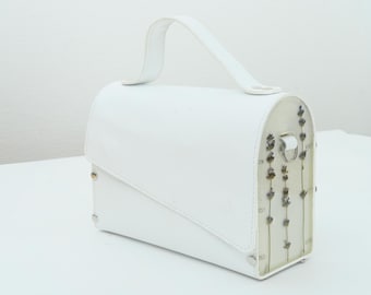 May include: White handbag with a curved top and a handle. The bag features a flap closure and decorative dried flowers encased on one side. The bag has a rectangular shape with rounded edges and metal accents.