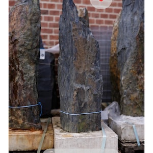 May include: A collection of large, dark gray and brown natural stone slabs. The stones are various heights and widths, with rough, textured surfaces. The dimensions of the stone are 116x33x11 cm. The stones are displayed on wooden pallets.