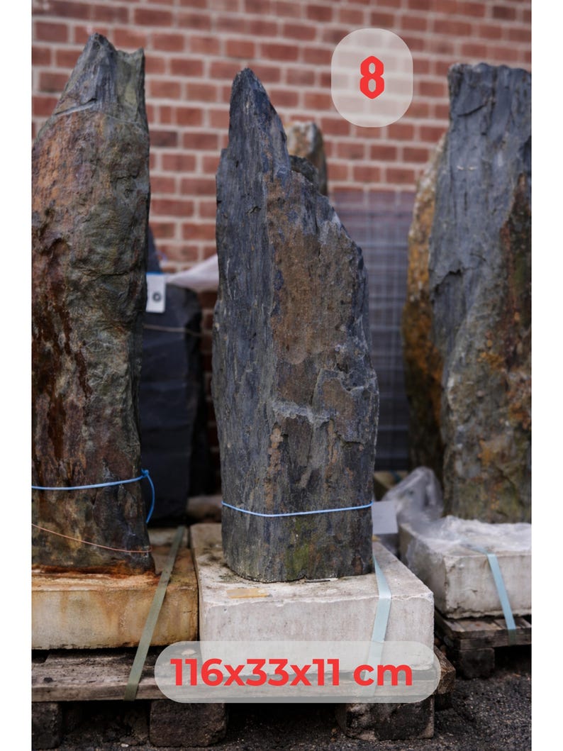 May include: A collection of large, dark gray and brown natural stone slabs. The stones are various heights and widths, with rough, textured surfaces. The dimensions of the stone are 116x33x11 cm. The stones are displayed on wooden pallets.
