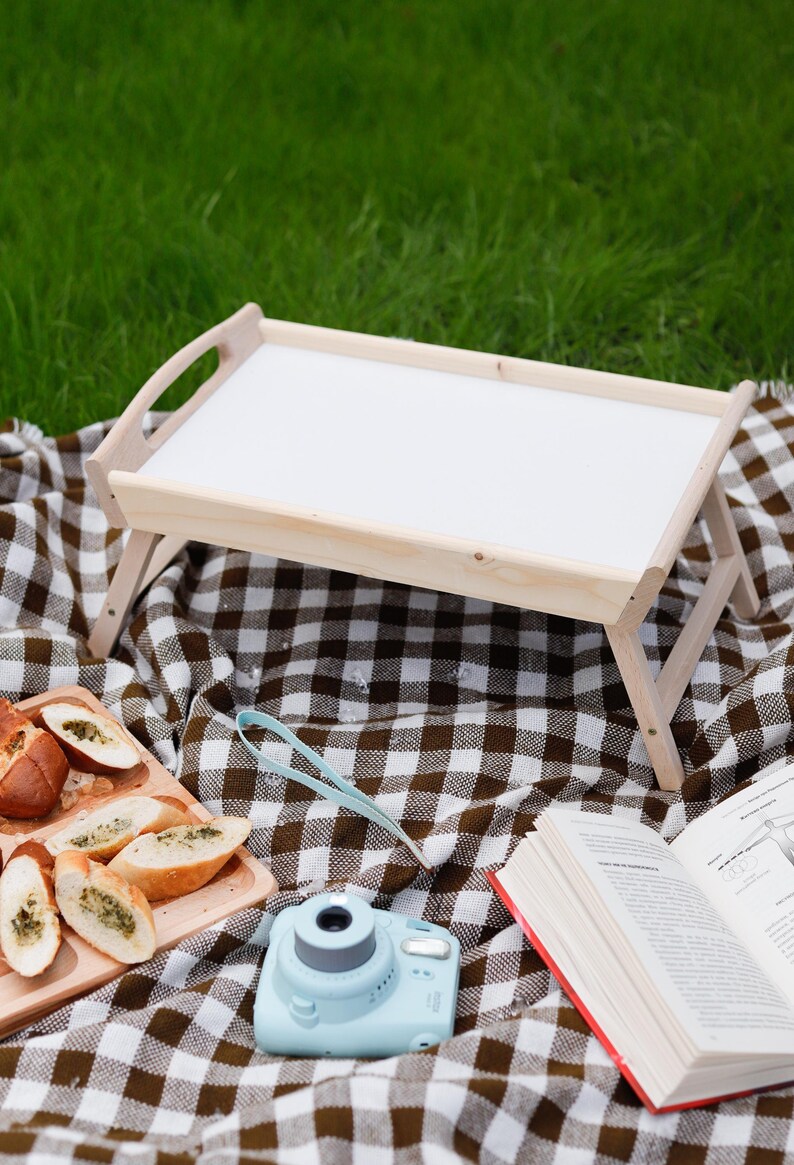 Klappbarer Betttisch mit weißer Tischplatte und Holzbeinen auf einer karierten Picknickdecke im Grünen.