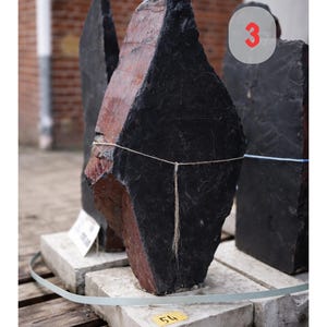 May include: A large, irregularly shaped black and red stone, possibly slate, with dimensions of 76x40x8 cm. The stone is displayed on a concrete base, with a blurred background of a brick building. The number "3" is visible.