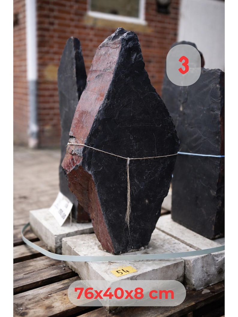 May include: A large, irregularly shaped black and red stone, possibly slate, with dimensions of 76x40x8 cm. The stone is displayed on a concrete base, with a blurred background of a brick building. The number "3" is visible.