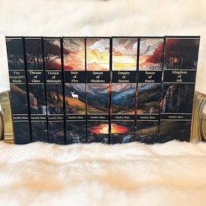 May include: A collection of fantasy novels, featuring titles like "Throne of Glass" and "Kingdom of Ash," displayed in a decorative gold-toned book holder. The book spines are black with landscape art and gold lettering. The author's name, Sarah J. Maas, is printed on each book.
