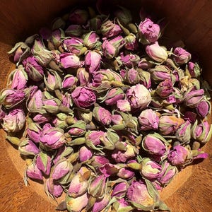 May include: A bowl filled with dried rose buds. The buds are a pale pink colour and have a green stem. The bowl is made of wood and has a natural finish.