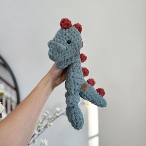 May include: A handmade, blue crochet dinosaur plush toy with red spikes and accents. The soft toy has a round, textured body and a small, round wooden tag. The dinosaur is held up against a white background.