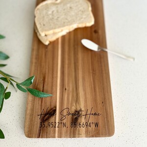 May include: A wooden chopping board with a handle, engraved with "Home Sweet Home" and coordinates. Two slices of bread and a butter knife are on the board. The board is made of light brown wood.