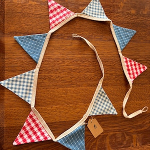 May include: A decorative bunting with alternating red and white gingham, blue and white check, and solid blue triangle flags. The flags are connected by a natural-colored fabric strip. A small, blank tag hangs from the end.