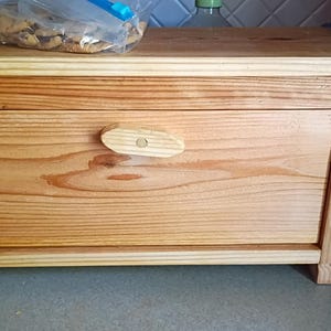 May include: A rectangular wooden bread box with a natural light brown wood grain finish, featuring a front-hinged door with a horizontal wooden handle and a flat top surface. The structure is constructed from light-colored pine-like wood planks.