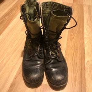 May include: A pair of worn black combat boots with olive green canvas tops and black laces. The boots are scuffed and show signs of wear, suggesting they have been used. The boots are placed on a wooden surface.