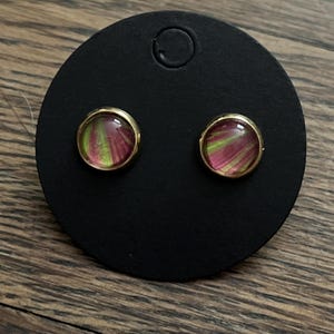 May include: A pair of stud earrings with a gold-colored bezel. The earrings feature a circular design with a pink and green abstract pattern. They are displayed on a black earring card.