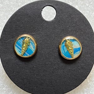 May include: A pair of gold-toned stud earrings. Each earring features a round, blue and white background with a gold-colored seashell design in the center. The earrings are displayed on a black card.