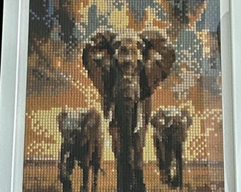 May include: Framed diamond painting of three elephants in a sunset scene. The artwork features a detailed mosaic of small, colorful squares, creating a textured image. The elephants are depicted in shades of brown and gray, set against a warm, golden sky.