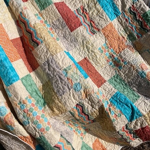以下が含まれることがあります： オレンジ、ターコイズ、グリーン、ブラウンの色合いの幾何学模様が様々なパッチワークのキルト。キルトはコットン生地で作られており、キルティングされた質感があります。