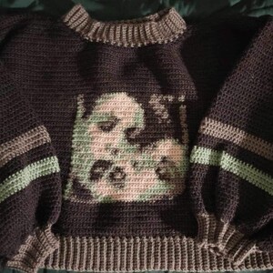 May include: A brown crocheted sweater with green and brown stripes on the sleeves. The sweater features a graphic of a face in the centre.