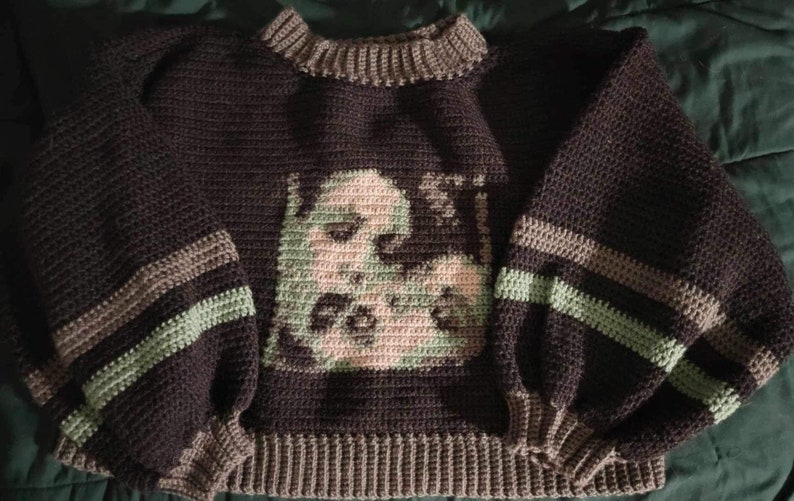 May include: A brown crocheted sweater with green and brown stripes on the sleeves. The sweater features a graphic of a face in the centre.