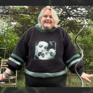 May include: A black and green crocheted jumper with a white graphic of a face. The jumper has green stripes on the sleeves.