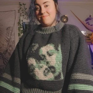 Może przedstawiać: Sweter zrobiony na szydełku w kolorze ciemnozielonym, szarym i jasnozielonym z graficznym wzorem twarzy. Sweter ma okrągły dekolt i długie rękawy.