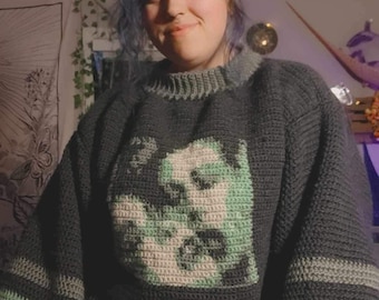 May include: A dark green, grey, and light green crocheted jumper with a graphic design of a face. The jumper has a crew neck and long sleeves.