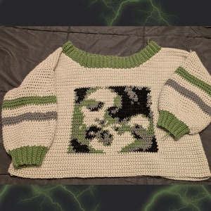 May include: A crocheted sweater with a green, black and grey geometric design on the front. The sweater has green and grey stripes on the sleeves and a green neckline.